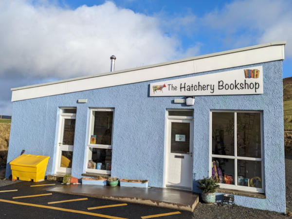 The front of the bookshop, with an angled roof, light blue textured render, two tall windows and two white doors. A sign reads 'The Hatchery Bookshop' and has a picture of a multicoloured cow and a row of colourful books.